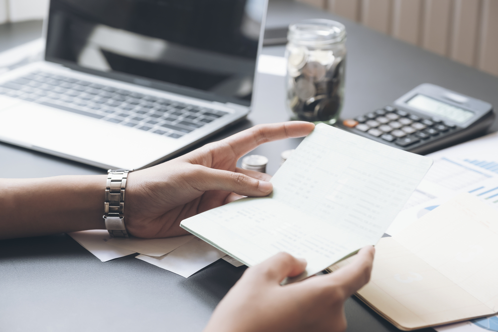 Photo of a person's hands holding a check register in front of a desk covered with papers, coins, a calculator and a computer.